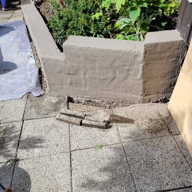 Nyputsad stödmur i en villaträdgård i Flen intill en stensatt uteplats och grönskande rabatt.