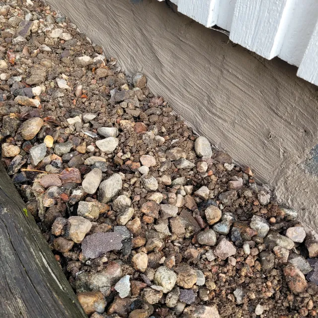 Närbild av stenig jord med stenar i olika storlekar intill en husgrund och vit träfasad. Till vänster syns kanten av en väderbiten träbalk.