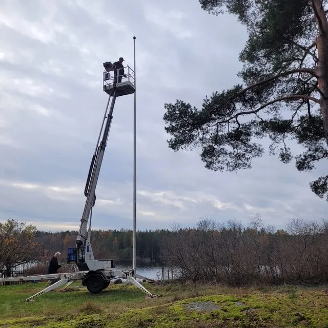 Två medarbetare från Boing All-Service i skylift vid en hög flaggstång nära en sjö vid byte av flagglina och rengöring.