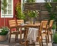 En matgrupp i trä med fyra stolar och ett bord är placerad utomhus. På bordet står en vas med blommor, tallrikar och tillbringare. En filt hänger på en stol. Gröna växter och ett rött trähus syns i bakgrunden.