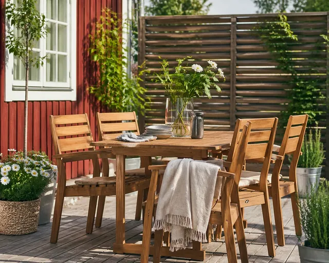 En matgrupp i trä med fyra stolar och ett bord är placerad utomhus. På bordet står en vas med blommor, tallrikar och tillbringare. En filt hänger på en stol. Gröna växter och ett rött trähus syns i bakgrunden.