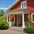 Välbyggd farstukvist i trä på ett rött svenskt trähus med vita lister. Blå himmel, sommardag.
