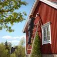 En målare stryker falu rödfärg på en träfasad en solig vårdag med stege vid husväggen.