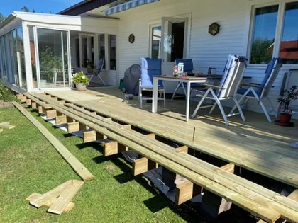 En uteplats på bakgården under uppbyggnad, med träbjälkar och plankor delvis installerade. Uteplatsen har utemöbler inklusive bord, stolar och växter. Ett vitt hus med stora fönster och ett inglasat solrum finns i bakgrunden.