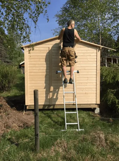En person i svart linne och bruna shorts står på en stege och hamrar eller arbetar på träpanelen i ett litet skjul. Boden ligger i ett gräsområde med träd och annan grönska runt omkring, under en klarblå himmel.