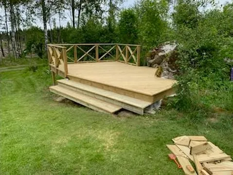 Ett nybyggt trädäck med trappor står på en gräsbevuxen bakgård. Däcket har ett räcke på två sidor och är omgivet av träd och grönt bladverk. En hög med träplankor och byggmaterial ligger på gräset till höger om däcket.