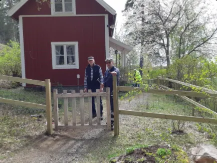 Två personer står framför en träport som leder till ett rött hus omgivet av grönska. En person lutar sig mot grinden, medan den andra står bredvid dem. Scenen verkar vara solig med träd och växter i bakgrunden.