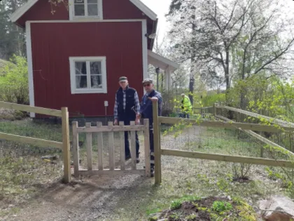 Två personer står framför en träport som leder till ett rött hus omgivet av grönska. En person lutar sig mot grinden, medan den andra står bredvid dem. Scenen verkar vara solig med träd och växter i bakgrunden.