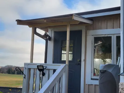 En liten veranda med takkonstruktion i trä och en mörkblå dörr leder in i ett beige hus. Verandan har vita räcken, en armatur och en metallstol. Bakgrunden visar ett öppet fält och en molnig himmel.