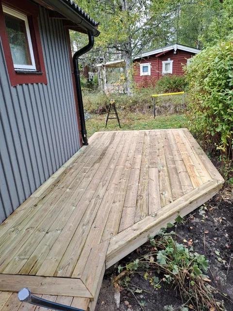 Ett nybyggt trädäck på ett grått hus med röda fasader, omgivet av grönska och buskar. Ett rött skjul syns i bakgrunden och verktyg står uppställda på gräsmattan.