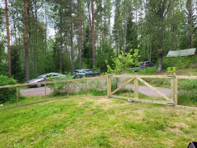 Ett trästaket med en öppen grind omger en gräsbevuxen gårdsplan. Bortom staketet står flera bilar parkerade på en grusad uppfart bland höga gröna träd i ett skogsområde.