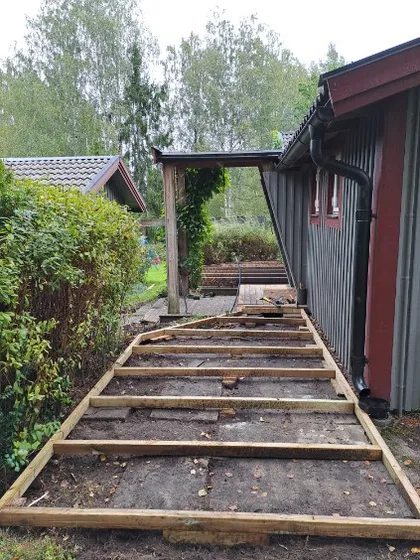 Träkonstruktion för ett däck eller en gångväg under uppförande intill ett hus, med jord och glest gräs mellan plankorna. Buskar kantar ena sidan och träd syns i bakgrunden.