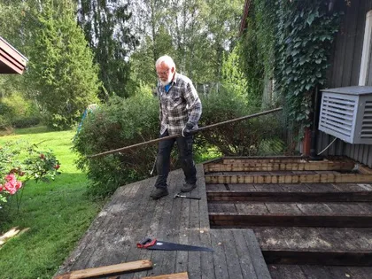 En äldre man i arbetskläder står på ett trädäck under reparation och håller i en lång metallstång. Verktyg och träplankor ligger på däcket, och gröna träd och buskar fyller bakgrunden en solig dag.