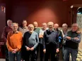 En grupp på tio äldre män står tätt tillsammans inomhus och ler mot kameran. De är vardagligt klädda och ser ut att befinna sig i ett varmt upplyst rum med en mörkröd vägg och ljus träpanel bakom dem.