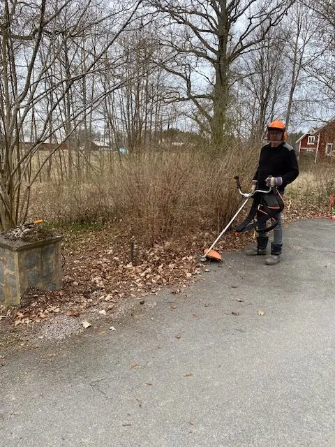 En person som bär säkerhetsutrustning använder en grästrimmer för att klippa gräs och ogräs längs en asfalterad gångväg.
