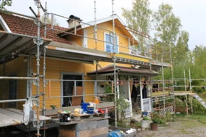 Ett gult trähus med rött tegeltak är omgivet av byggnadsställningar och byggmaterial, vilket tyder på pågående renoveringar. Verandan har olika föremål, inklusive en stege, verktyg och krukväxter. Träd och buskar är i bakgrunden.