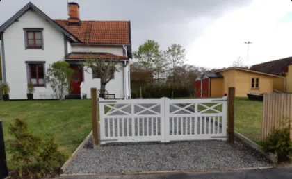 Ett vitt hus med rött tegeltak står bakom ett vitt staket med dubbla grindar. Huset har röda fönsterkarmar, en liten veranda och en trimmad trädgård med ett träd. Ett träskjul syns till höger, och himlen är molnig.