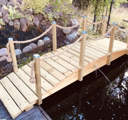 En liten träbro med räcken spänner över en lugn damm. Bron, konstruerad av ljust trä, har robusta stolpar förbundna med tjocka, knutna rep. I bakgrunden finns klippor och grönska som omger vattnet.