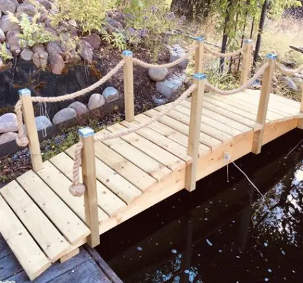 En liten träbro med räcken spänner över en lugn damm. Bron, konstruerad av ljust trä, har robusta stolpar förbundna med tjocka, knutna rep. I bakgrunden finns klippor och grönska som omger vattnet.