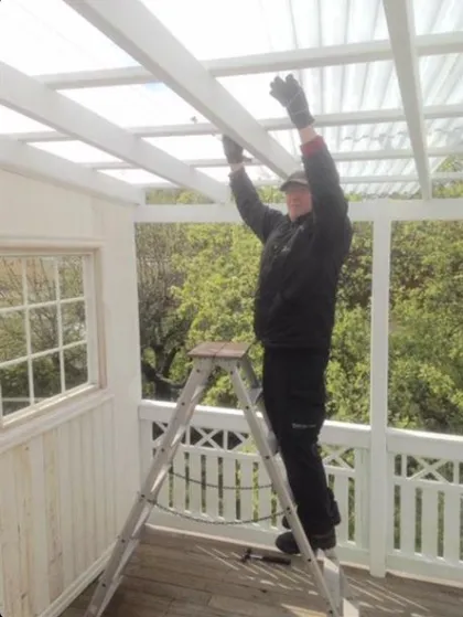 En person i svart jacka och handskar står på en stege och arbetar på taket av en vit träpergola. Pergolan har utsikt över en naturskön utsikt med träd och ett delvis vitt staket som syns nedanför.