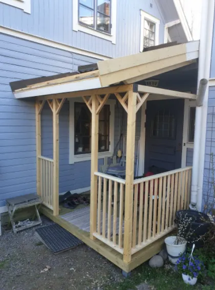 En ofullbordad veranda i trä med delvis färdigbyggt tak fäst vid ett grått hus. Verandan har vertikala räcken och en liten uppsättning trappsteg som leder upp till entrén. Olika föremål, inklusive skor och krukväxter, placeras runt verandan.