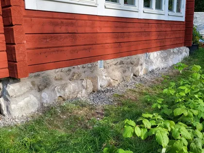 Närbild på en renoverad stenmur och husgrund med murbruk och ventilationsöppningar på ett rött trähus.