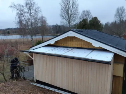 Ett litet träskjul med svart tak står nära en sjö omgiven av kala träd. En person arbetar på vänster sida av boden, eventuellt med bygg- eller underhållsarbete. Marken är täckt av löv och himlen verkar mulen.