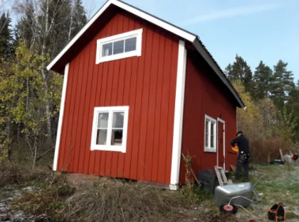 En person arbetar på utsidan av ett litet tvåvånings rött trähus med vita detaljer, beläget i ett lantligt område omgivet av träd. Marken verkar frostig och det finns verktyg och utrustning utspridda nära byggnaden.
