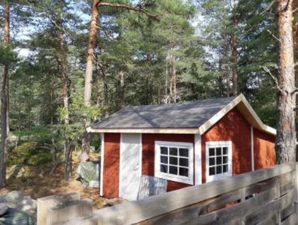 En pittoresk röd trästuga med vita detaljer ligger inbäddat bland höga tallar. Det finns ett trästaket i förgrunden, och stugans entré har en vit dörr och ett fönster med flera rutor. Omgivningen är frodig med grönt lövverk.