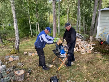 Två personer använder en vedklyv utomhus i ett skogsområde. En person använder maskinen medan den andra placerar en stock. Olika hackade träbitar är utspridda runt dem. I bakgrunden finns en hög med stockar och vad som verkar vara en stuga eller ett skjul.