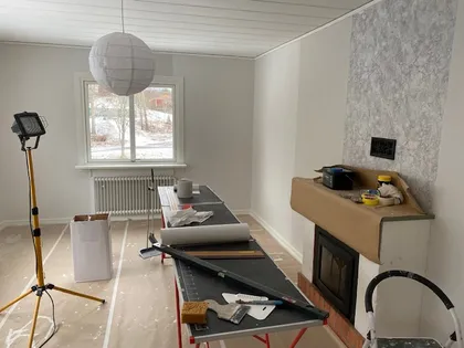 Ett rum som håller på att renoveras. Golvet är täckt med skyddande lakan och olika renoveringsverktyg och material finns på hopfällbara bord. En öppen spis är delvis avtejpad för skydd och ett stort fönster släpper in naturligt ljus i utrymmet.