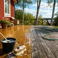 Ett altandäck som tvättas med såpa och borste. Ena halvan är ren och ljus, den andra fortfarande grå. Röd stuga i bakgrunden.