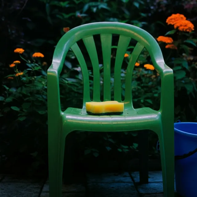 En grön plaststol med en gul svamp på sitsen står på en uteplats, omgiven av blommande orange ringblommor och grönska. En blå hink är placerad på marken bredvid stolen.