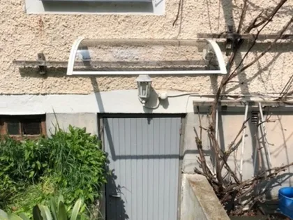 En väderbiten, beige stuckatur byggnadsvägg med en liten, grå metalldörr och ett genomskinligt, svängt takskydd ovanför. Till vänster om dörren spirar gröna växter och till höger bladlösa grenar. En vit armatur är monterad ovanför dörren.