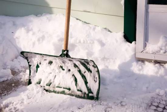 En stor grön snöskyffel med trähandtag lutar sig mot en snöhög intill ett hus i Katrineholm.